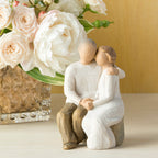 Willow Tree Anniversary, 6”h hand-painted resin figure of older couple sitting on bench holding hands, both in neutral clothing, on table with white roses