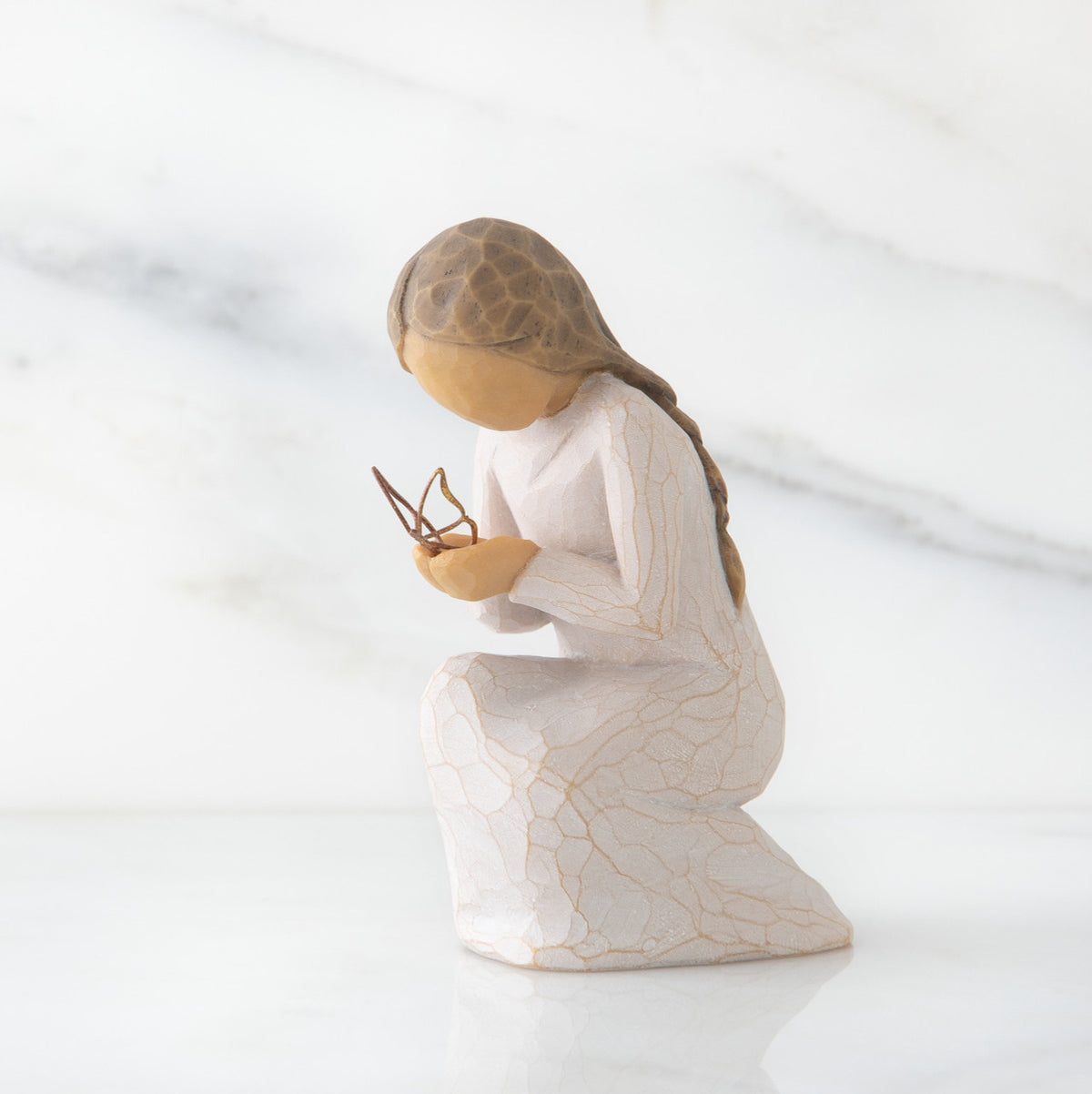 Side view of kneeling female figure in cream dress, with wire butterfly on extended hands, on marble background