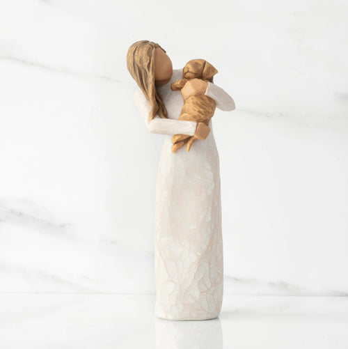 Front view, standing female figure in cream dress, holding golden color puppy in her arms