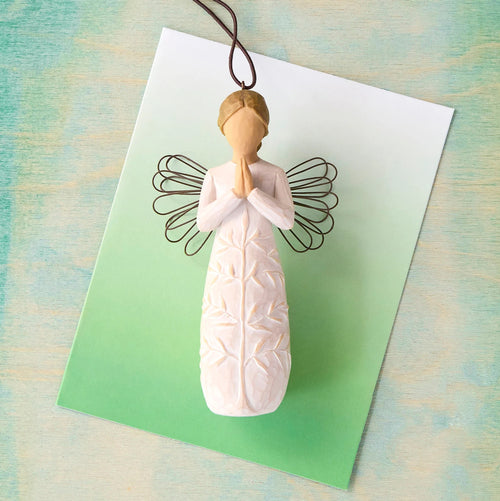 Willow Tree a tree, a prayer Ornament, 4”h hand-painted resin angel figure in cream dress with wire wings, hands in prayer position, tree carving on dress, brown elastic cord, laying in front of green painting