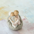 Willow Tree My new baby (blush), 2.5”h hand-painted resin figure of toddler in blush pink onesie holding infant in cream, cuddled together in a beanbag, in front of pastel painting