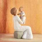 Willow Tree Grandmother, 5.5”h hand-painted resin figure of seated older woman in cream dress, holding baby up in the air, in front of orange painting