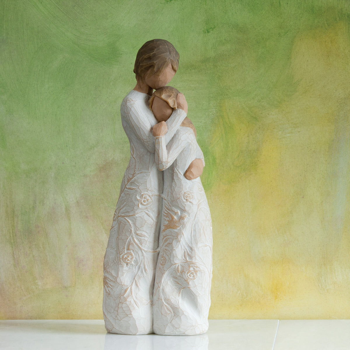 Willow Tree Close to me, 8"h hand-painted standing resin figure of woman embracing younger girl, both in cream dresses carved with intertwined vines and leaves, in front of olive green painting
