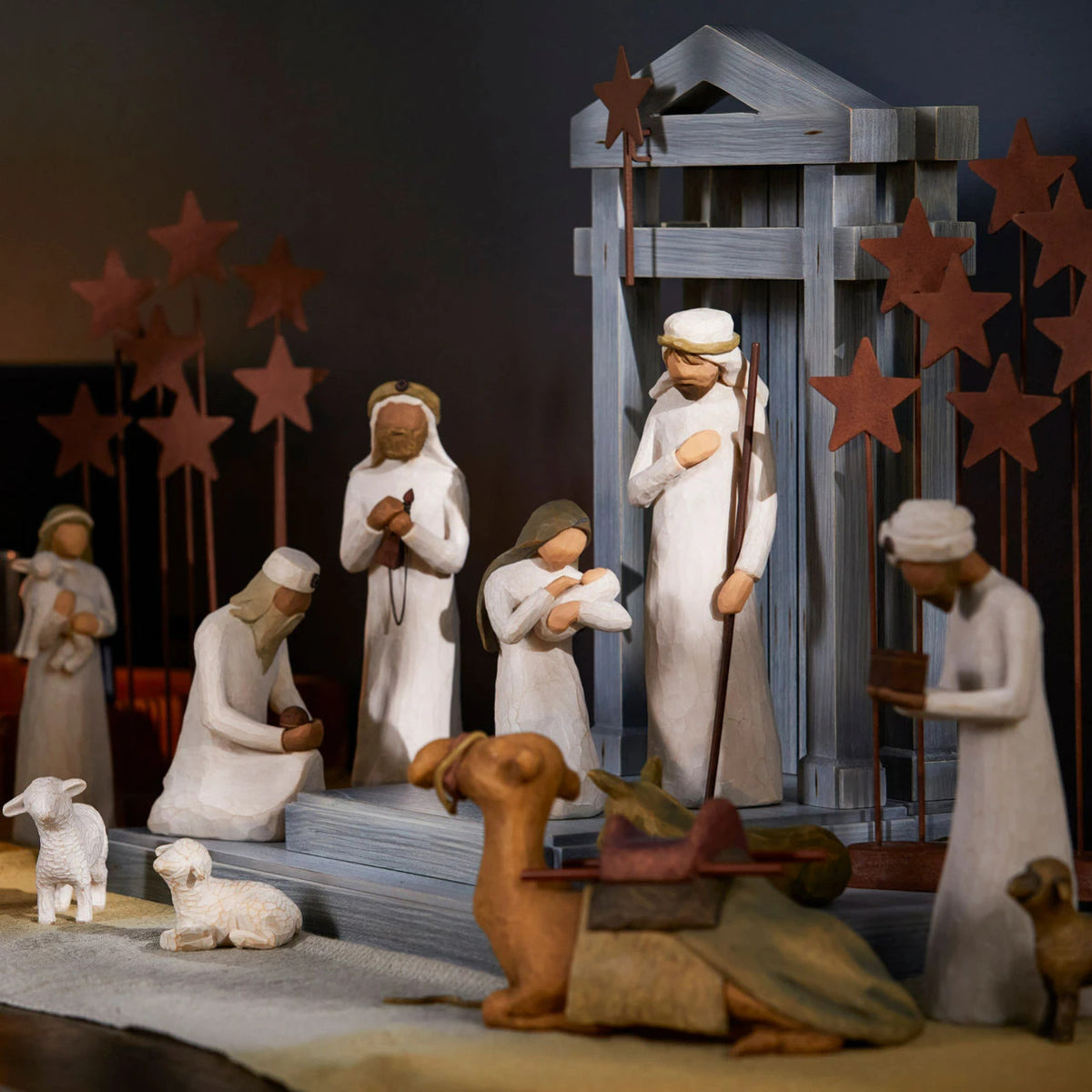 A carved nativity scene set out on a wood table laid with a linen cloth and lit candles in the background.