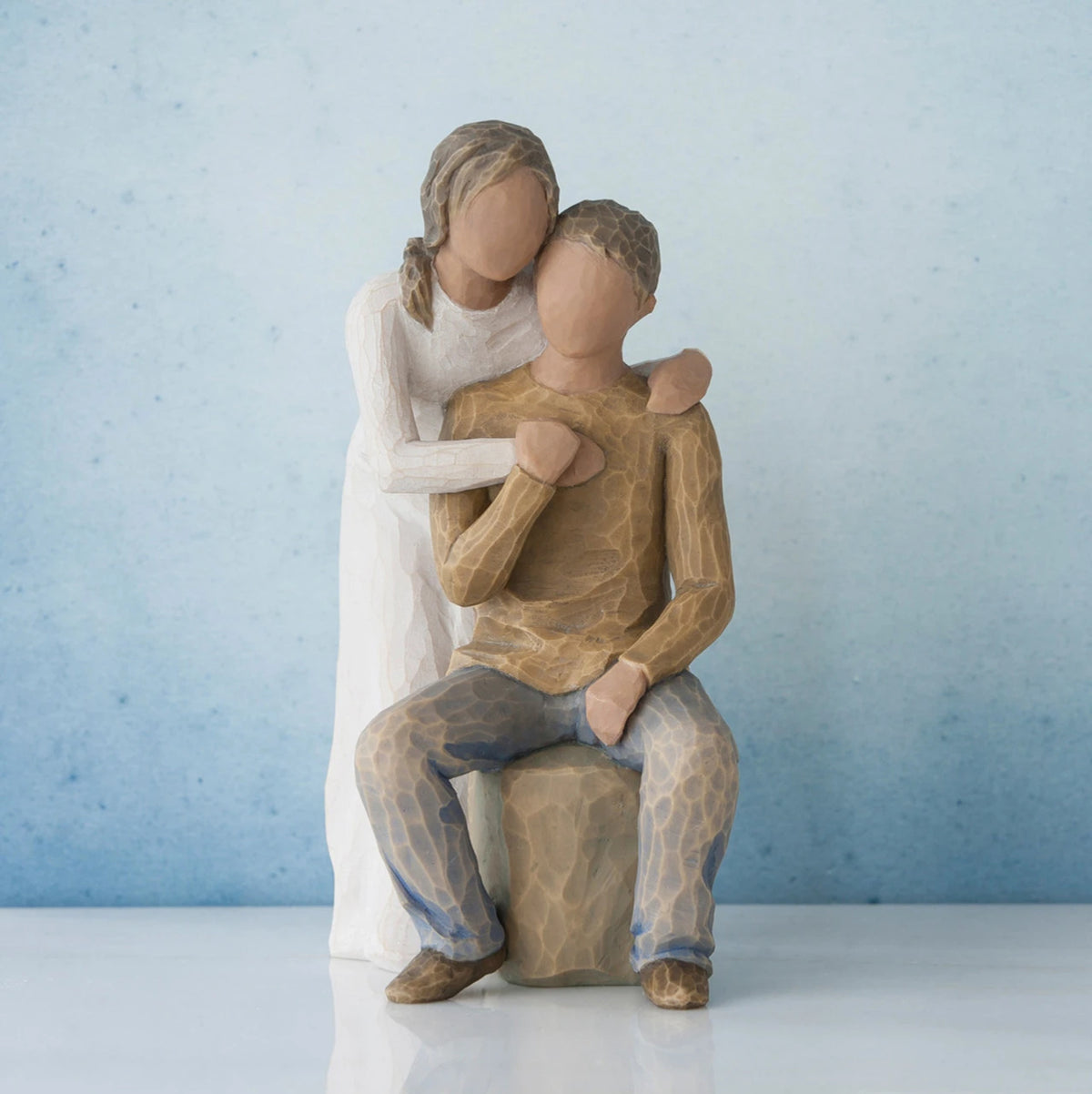 Man figurine sitting on rock wearing brown shirt and blue jeans with woman figurine in white dress with arms wrapped around him - blue background