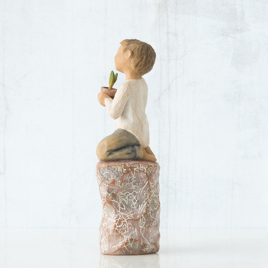Right side view of boy with brown hair sitting on a stump while holding a small potted plant to his chest