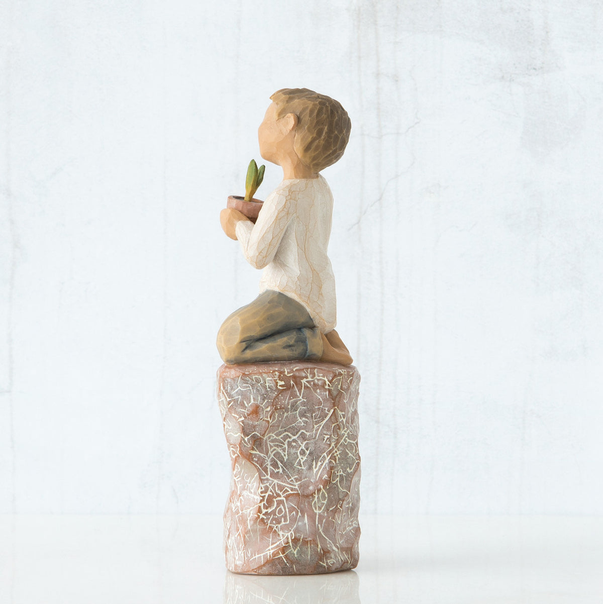 Right side view of boy with brown hair sitting on a stump while holding a small potted plant to his chest