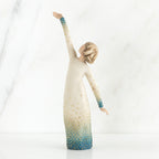 Faceless woman figurine in white dress with blue embellishments - she is facing away from the camera holding both her arms in opposite directions