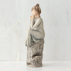 Brunette girl figurine in light grey dress sitting on grey rock facing the side - white background