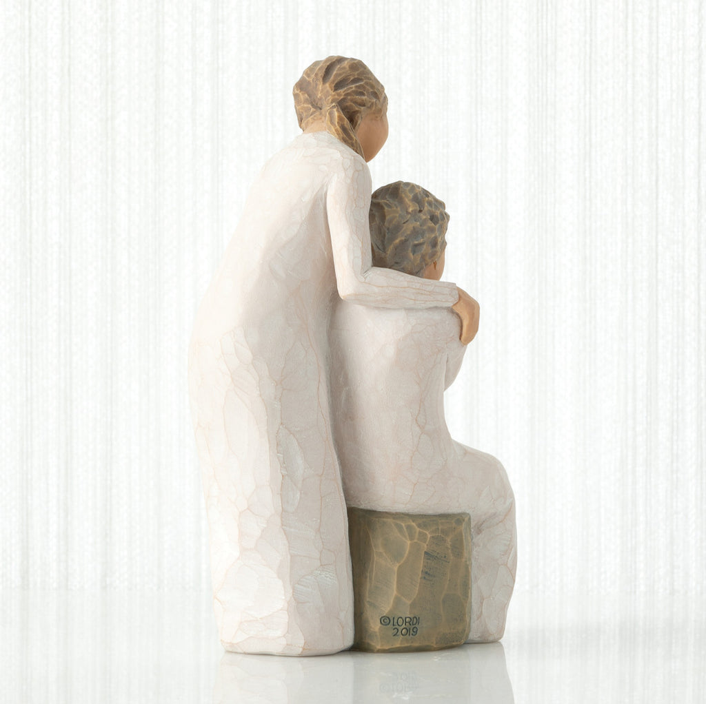 Back view, seated older female in cream dress, with younger female in cream dress, standing at her shoulder, hands embracing