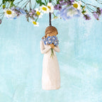Willow Tree Forget-me-not Ornament, 4.25”h hand-painted resin figure in cream dress, holding bouquet blue flowers up to face, brown elastic cord, hanging in front of blue painting