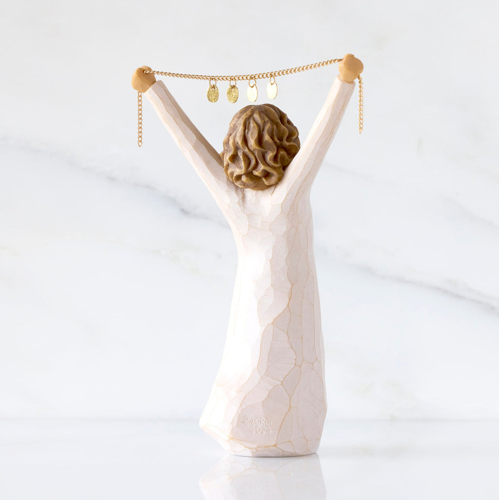 Willow Tree Courage 2025, back view of 6”h hand-painted standing figure in cream dress holding aloft a gold chain with 2025 etched on gold medallions