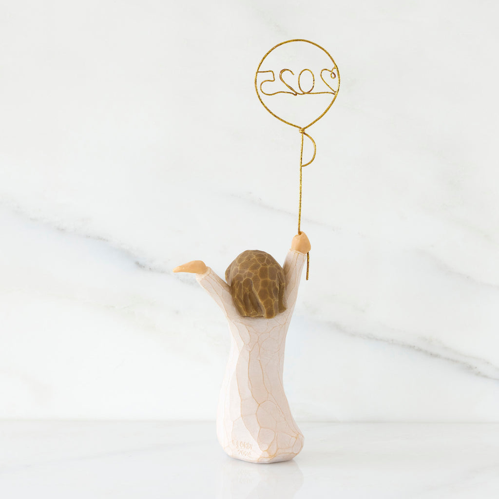 Back view: standing figure in cream dress, arms raised, holding gold wire balloon with 2025 written in gold wire inside
