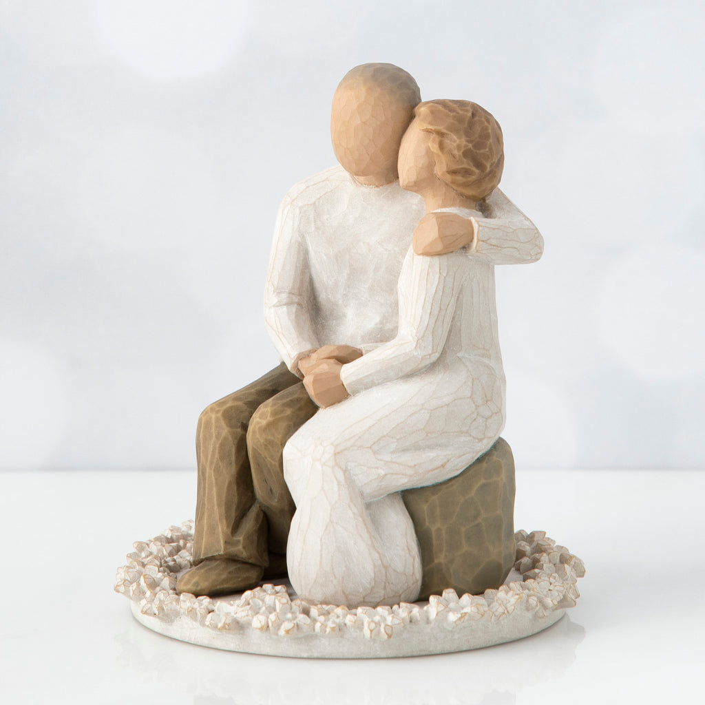 Man and woman figurine in white sitting on a rock on top of round white plaque hugging one another