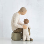 Father figurine in brown pants and white shirt sitting on rock with arm around little boy figurine in white