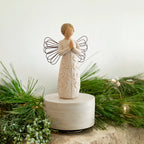 Willow Tree a tree, a prayer Musical, 7.5”h hand-painted resin angel figure in cream dress with wire wings, hands in prayer position, tree carving on dress, atop round, cream base containing musical element, on table with fir branches
