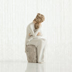 Woman figurine sitting on grey rock dressed in white holding small baby facing away from the front - white background