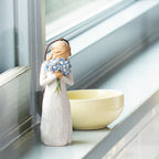 Willow Tree Forget-me-not Ornament, 4.25”h hand-painted resin figure in cream dress, holding bouquet blue flowers up to face, brown elastic cord, on bright windowsil