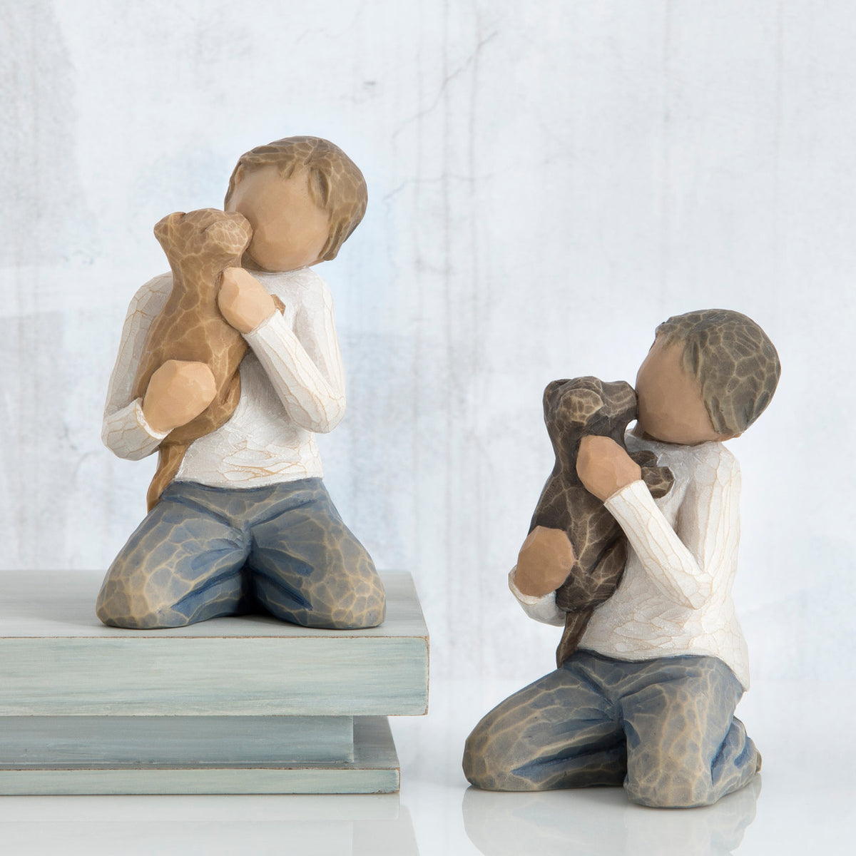 Front view of boy with light brown hair wearing white shirt and jeans and holding a light brown puppy as it licks his face. Beside him, front view of boy with dark brown hair wearing white shirt and jeans and holding a dark brown puppy as it licks his face.