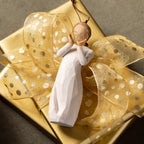 Willow Tree Lots of Love Ornament, 4.5”h hand-painted resin figure in cream dress, holding wire concentric heart with gold glitter, brown elastic cord, tied to gold gift