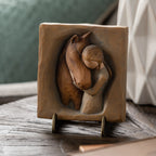 Willow Tree Hug Plaque, 4”sq x .5”d hand-painted resin bas-relief carving of girl in cream dress, arms around neck of reddish-color horse, on green/gray background. Plaque displayed on Plaque Stand on table