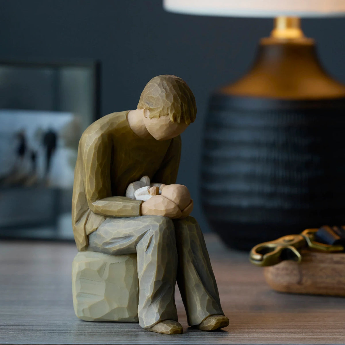 Willow Tree New Dad, 6”h hand-painted seated resin figure in cream shirt and jeans, gazing at newborn infant on lap, on shelf with lamp