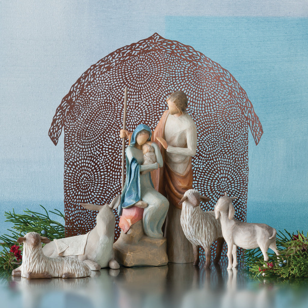 Nativity scene figurines sitting down holding baby next to lambs with brown barn in the background