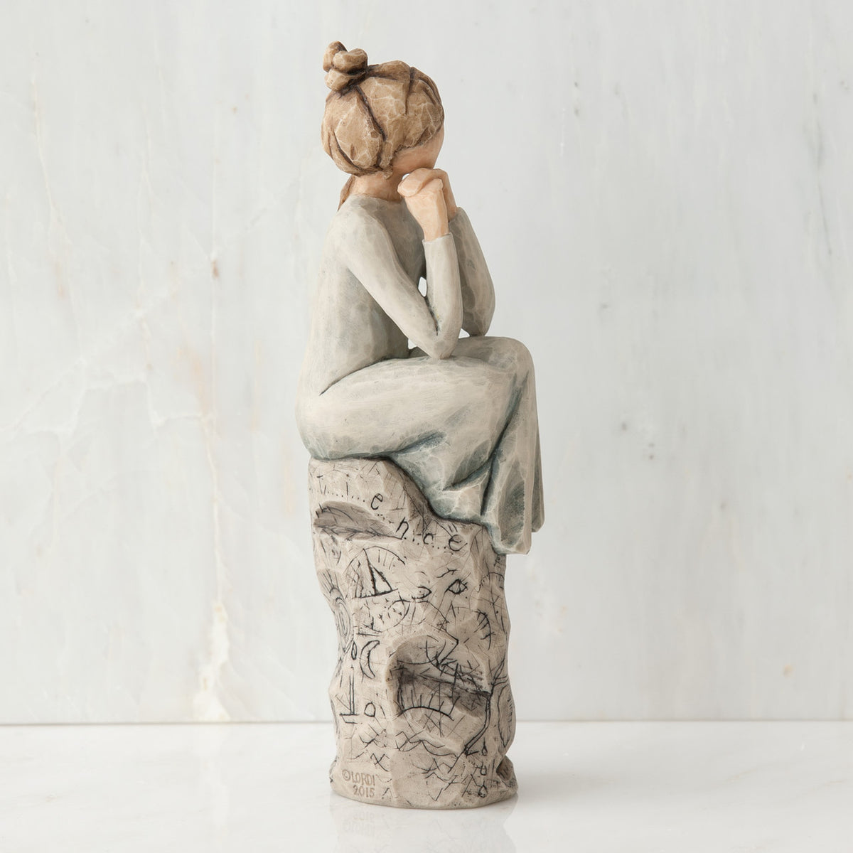 Brunette girl figurine sitting on grey  rock in grey dress with her hands up to her face