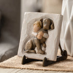 Willow Tree Hug Plaque, 4”sq x .5”d hand-painted resin bas-relief carving of small boy in cream shirt, hugging large dark dog. Plaque displayed on Plaque Stand on table