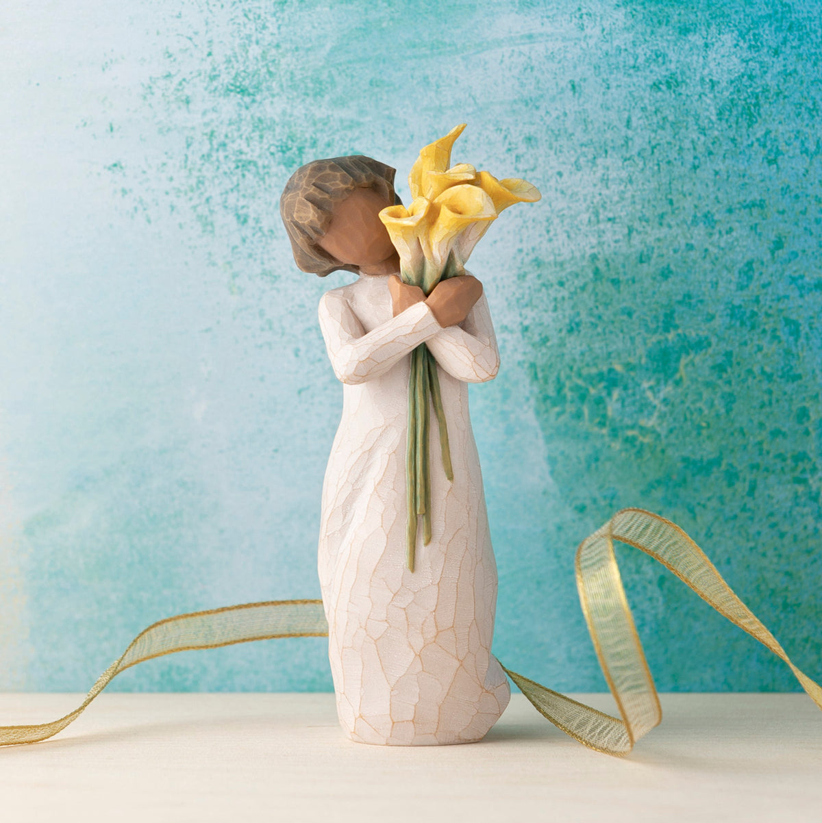 Willow Tree With Gratitude, 5.5”h hand-painted resin figure with darker skin and hair, in cream dress, standing, holding bouquet of yellow calla lilies, in front of blue painting
