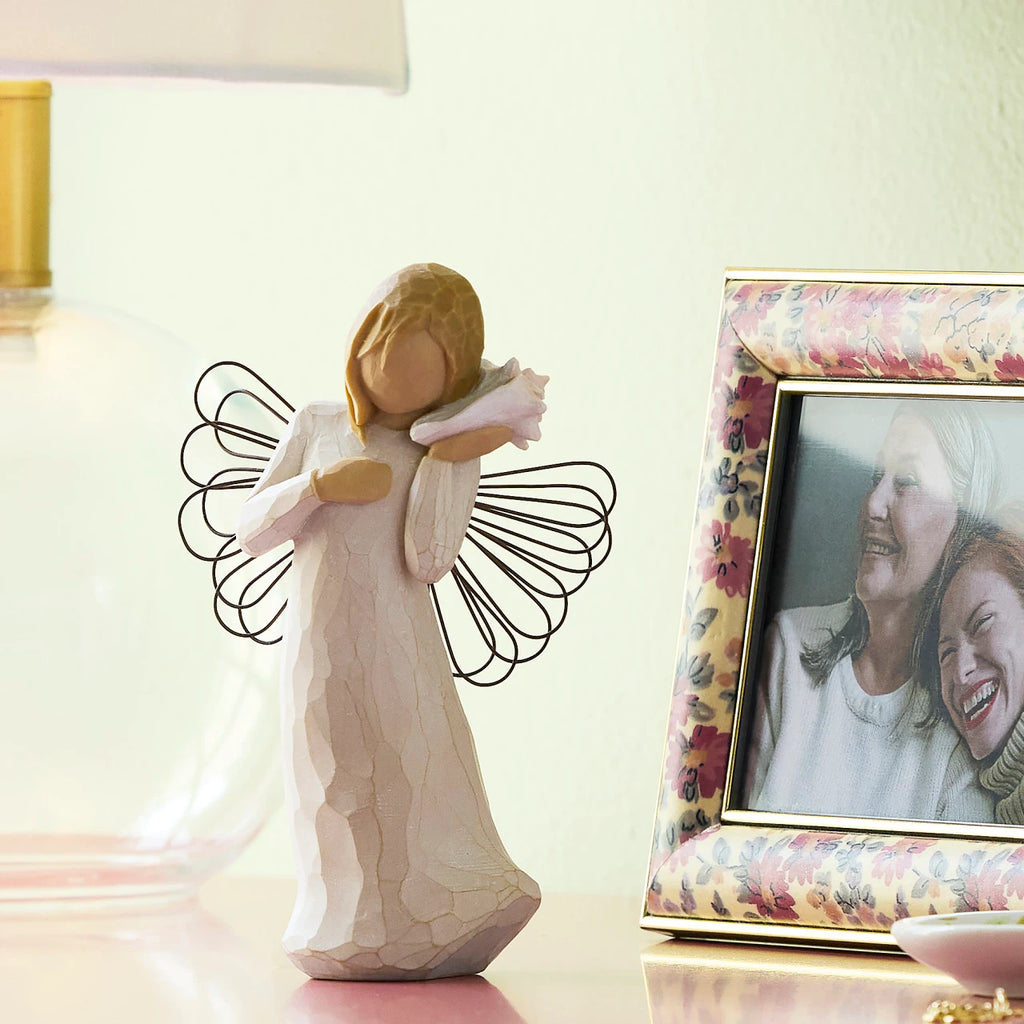 Willow Tree Thinking of You, 5.5”h hand-painted standing resin angel figure in cream dress with wire wings, holding pink conch shell to ear, on shelf with photos