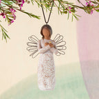 Willow Tree Remembrance Ornament, 4”h hand-painted resin angel figure with darker skin and hair, wire wings, arms crossed over gold heart, cream dress carved with nature imagery, brown elastic cord, hanging in front of pastel painting