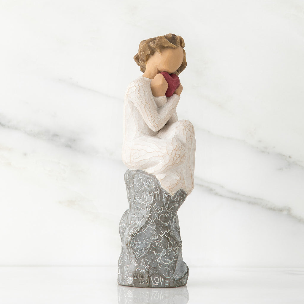 Side view of girl figurine in white sitting on grey rock holding pink heart figurine