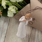 Willow Tree Thinking of You Ornament, 4.5”h hand-painted resin angel figure in cream dress with wire wings, holding pink conch shell to ear, brown elastic cord, laying next to floral bouquet