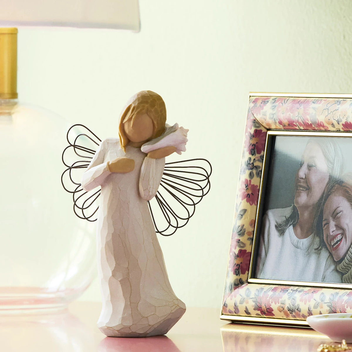 Willow Tree Thinking of You, 5.5”h hand-painted standing resin angel figure in cream dress with wire wings, holding pink conch shell to ear, on shelf with photos