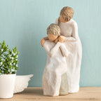 Willow Tree Loving My Mother, 6.5”h hand-painted resin figure of seated older woman with younger woman standing at her shoulder, both in cream dresses, hands clasped, in front of muted teal painting