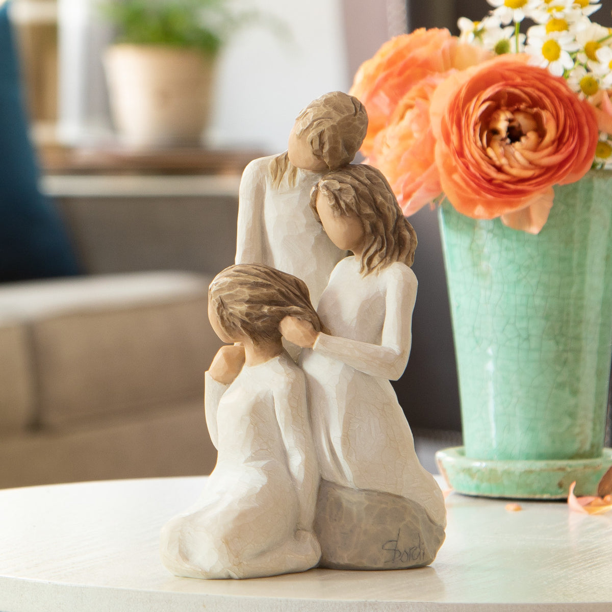 Willow Tree Our healing touch, 6.5”h hand-painted resin figure of three women; one standing with arm around kneeling girl, holding hand of seated girl, kneeling girl arranging hair of seated girl, all in cream dresses, on table with coral flowers