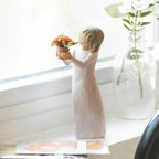 Willow Tree Little Things, 5.25”h hand-painted standing resin figure in cream dress, holding out terracotta pot with orange flowers, on bright windowsill
