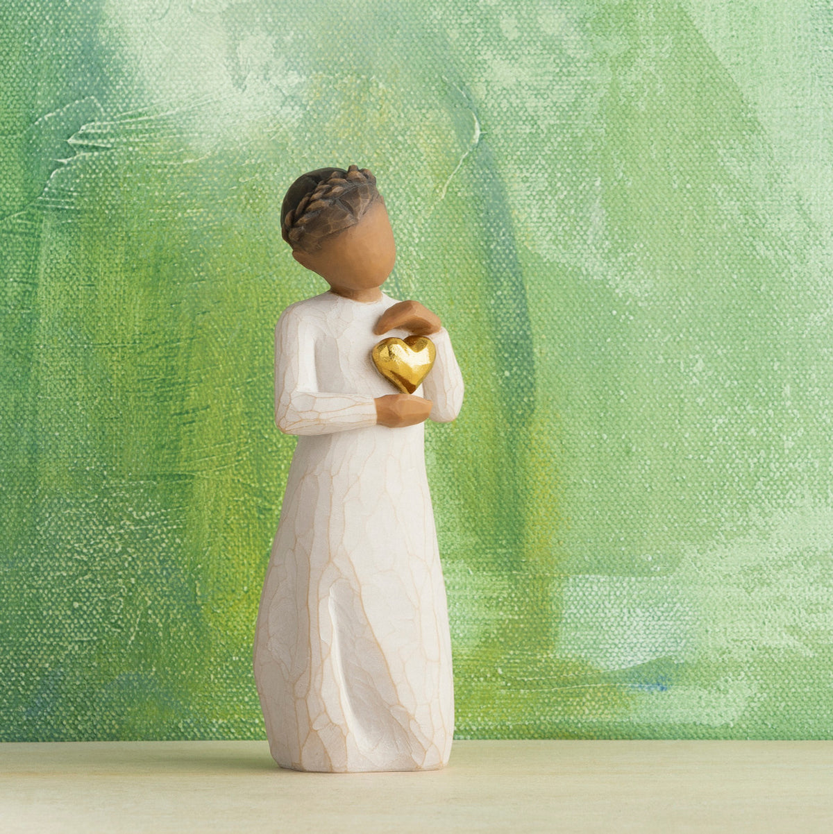 Willow Tree Keepsake, 5.5”h hand-painted standing resin figure with darker skin and hair, in cream dress, framing gold heart with both hands on her chest, in front of green painting